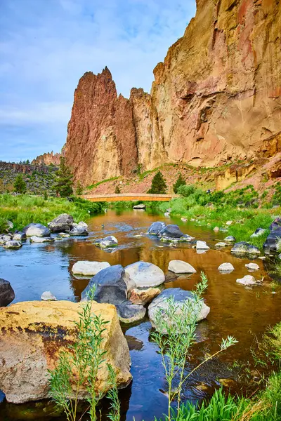 stock image Majestic cliffs at Smith Rock State Park in Oregon tower over a serene river and rustic wooden bridge, surrounded by lush greenery. Perfect for travel, outdoor adventure, and nature-themed projects.