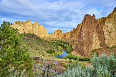 Oregon 'daki Majestic Smith Rock State Park' ta engebeli turuncu ve kahverengi uçurumlar, gökyüzünü yansıtan sakin bir nehir ve büyüleyici tahta bir yaya köprüsü bulunur. Seyahat ve doğa meraklıları için mükemmel.