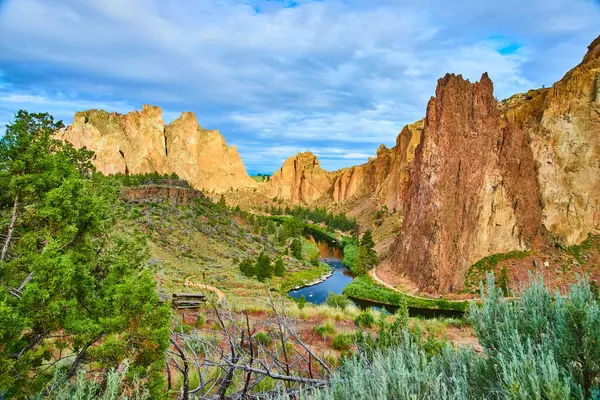 stock image Majestic Smith Rock State Park in Oregon features rugged orange and brown cliffs, a serene river mirroring the sky, and a charming wooden footbridge. Perfect for travel and nature enthusiasts seeking