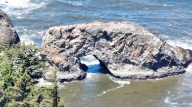 Oregon 'daki Samuel H. Boardman State Sahne Koridoru' ndaki Arch Rock 'ın hava görüntüsü turkuaz okyanus dalgaları arasında parlak güneş ışığı ve yemyeşil ağaçlarla vurgulanmış engebeli doğal kemeri yakalar.