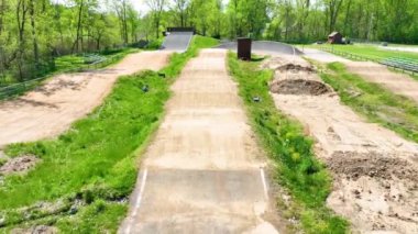 Varşova, Indiana 'daki bir BMX pistinin büyüleyici bir şekilde uçuşu, toprak izleri, rampalar ve yemyeşil alanları gösteriyor. Gün ortası aydınlatması BMX sürüşünün heyecanını vurguluyor