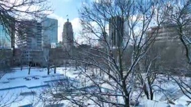 Fort Wayne 'deki Friemann Meydanı' nın sakin kış manzarası, karla kaplı ağaçlar, Allen County Adliyesi ve modern şehir binaları, bir karın barışçıl güzelliğini yansıtıyor.