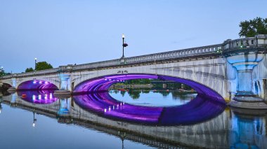 South Bend, Indiana 'da alacakaranlıkta güzel aydınlatılmış bir köprü. Canlı mor ve mavi ışıklar, River Lights Plaza 'nın sakin sularından yansıyarak tarihi kemerleri aydınlatıyor. Şehir zarafeti