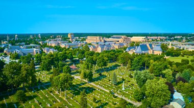 Aerial view of the University of Notre Dame campus in South Bend, Indiana, featuring historic and modern buildings amidst lush greenery, with a serene cemetery in the foreground under clear blue skies clipart