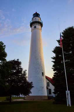 Golden hour at Wind Point Lighthouse in Wisconsin. This serene coastal landmark with its traditional design, red-roofed building, and gently fluttering American flag captures the essence of maritime clipart