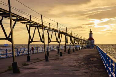 Michigan Gölü 'ndeki Benton Limanı Kuzey İskelesi' nde altın saat. Huzurlu bir günbatımı ve ikonik beyaz-kırmızı iç deniz feneri. Mavi parmaklıklarla süslenmiş sakin sahne, huzur yayıyor.