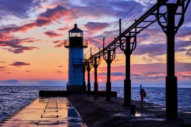 Benton Limanı Dış Deniz feneri gün batımında Michigan Gölü 'nde dimdik ayakta duruyor. Altın saatin ılık renkleri, nemli iskele yüzeyindeki güzellik ve sükuneti yansıtan sakin bir sahneyi resmeder.