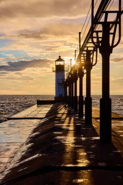 Benton Harbor, Michigan 'daki St. Joseph North Pier Deniz Feneri' nde altın saat. Batan güneş, ıslak iskele üzerinde sıcak bir parıltı yayar ve gemilere güvenli bir şekilde kıyıya ulaşmalarını sağlar. Huzurlu bir kılavuz ve...