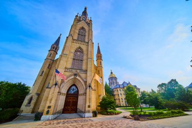 Majestic Gothic Revival facade of Notre Dame Basilica of the Sacred Heart in South Bend, Indiana. Captured from a low angle, the image features lush greenery, American pride, and a golden-domed clipart