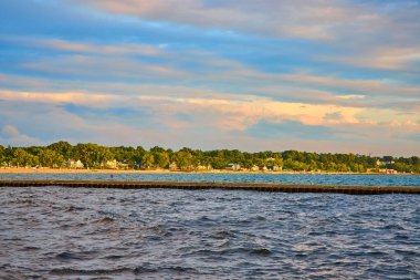 Michigan Gölü 'ndeki Benton Limanı' nda gün batımı, sakin bir kıyı sahnesi, hafif dalgalar, pitoresk kıyı şeridi ve büyüleyici evler sergiliyor. Rahatlama, turizm ve rıhtım konuları için mükemmel.