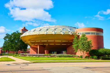 Modern red brick building with a striking metal-domed roof, surrounded by manicured lawns and trees, stands against a clear blue sky at Frank Lloyd Wrights SC Johnson Global Headquarters in Racine