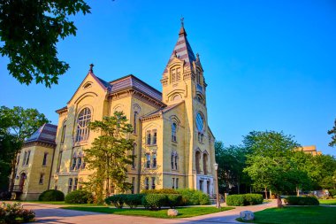 South Bend 'deki Notre Dame kampüsündeki görkemli tarihi bina altın ışıkla yıkanmış. Uzun kule, kemerli pencereler ve yemyeşil ile çevrili karmaşık taş işçiliği. Eğitim için mükemmel.