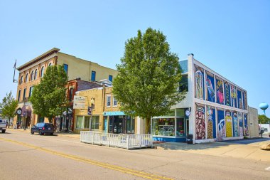 Charming Michigan main street in Three Oaks with colorful storefronts. Highlighted by a vibrant mural and the beloved Sweet on Elm candy shop. Trees and varied architecture add to the inviting scene.