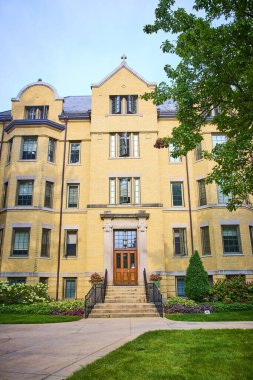 South Bend 'deki Notre Dame binasının görkemli sarı cephesi, parlak mavi gökyüzü altında taş sütunlar ve çiçek açan çiçek yatakları, akademik mükemmelliği ve tarihsel mükemmelliği somutlaştırıyor.