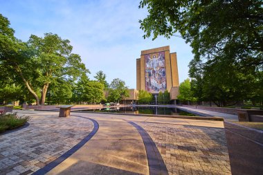 Notre Dame Hesburgh Kütüphanesi 'nde ikonik Hayat Sözcüğü duvar resmi İsa' nın dokunuşuyla. Yeşillik ve dingin bir yansıtma havuzu bu tarihi Indiana kampüsünde sakin bir sahne oluşturur..