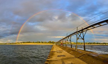 Golden hour serenity at North Pier, Benton Harbor, Michigan. A broad concrete pier leads into tranquil Lake Michigan waters, illuminated by a stunning double rainbow arching over the vibrant coastal clipart