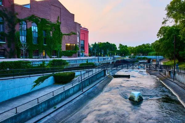 South Bends 'in pitoresk kanalında altın saat. Sarmaşık kaplı bir tuğla bina ve yaya köprüsü, doğa ve şehir hayatının huzurlu bir karışımını geliştirerek, iyi korunmuş bir kentsel sakinliği yansıtıyor.