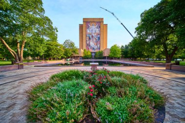 Serene morning at the University of Notre Dame, featuring the iconic Word of Life Mural on Hesburgh Library. Vibrant flowers, tranquil reflecting pool, and dynamic crane set a harmonious academic clipart