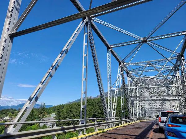 stock image Stunning steel truss bridge in Oregons mountains showcasing intricate engineering. Vehicles crossing under clear blue skies highlight day travel against lush green trees and scenic peaks.