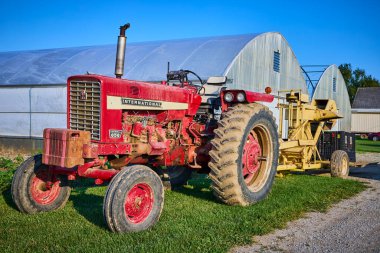 Klasik bir kırmızı Farmall 656 traktörü yeşil çimlerin üzerinde duruyor açık mavi Indiana gökyüzünün altındaki iki büyük seranın önünde geleneksel çiftlik hayatını modern tarım hayatıyla harmanlıyor.