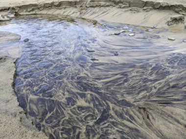 Oregon 'daki kumlu plajın üzerinden kristal berrak akıntı akar, ışık ve kum tanelerindeki büyüleyici desenleri ortaya çıkarır. Bu kıyı manzarasının doğal sükunetini ve güzelliğini hissedin..