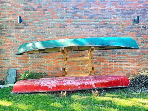 stock image Worn green and red canoes rest in a grassy Racine, Wisconsin backyard against a textured brick wall. Outdoor lighting enhances the rustic charm, evoking memories of past outdoor adventures.