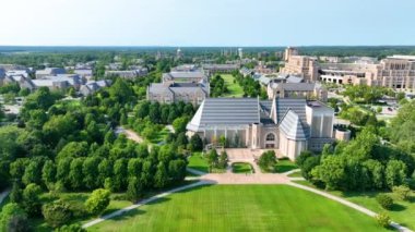 Aerial Fly Over Forward: The University of Notre Dame kampüsü South Bend, Indiana 'da ikonik binalar, yemyeşil alanlar ve parlak gündüz ışığı altında Altın Kubbe sergileniyor.