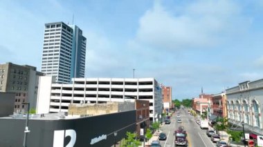 Pan Shot: South Bend Indiana şehir merkezinin havadan görünüşü, açık mavi gökyüzü altında tarihi ve modern mimarinin karışımını vurguluyor. Hareketli sokak manzarası zarif Palais Royale 'i sergiliyor.