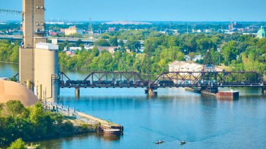 Toledo Ohio 'daki sakin Maumee Nehri üzerindeki paslı demiryolu köprüsünden geçen bir trenin hava görüntüsü. Endüstriyel silolar ve yemyeşil ağaçlar, huzurlu konutların zeminiyle tezat oluşturuyorlar..