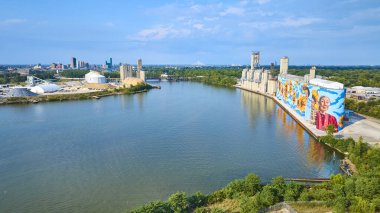 Aerial view of Toledo Glass City River Wall murals on silos depicting nature. Vibrant art contrasts the bustling Maumee Riverside industrial hub, reflecting community spirit and urban resilience. clipart