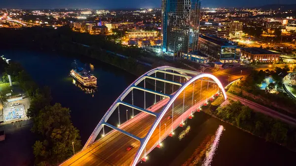 stock image Aerial view of Nashville at night with the Korean Veterans Memorial Bridge glowing over the serene Cumberland River. Downtowns illuminated skyline and a lit riverboat paint a vibrant urban scene.