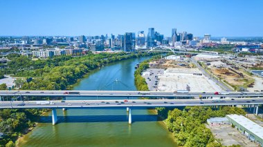 Nashville Tennessee 'nin havadan görünüşü açık mavi gökyüzü altında canlı bir şehir silueti, sakin Cumberland Nehri ve hareketli bir karayolu köprüsü ve kentsel büyüme ve doğal uyumu gösteriyor..