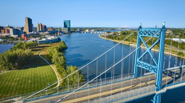 Toledo, Ohio 'daki Anthony Wayne Köprüsü' nün Maumee Nehri 'ni kaplayan havadan görüntüsü. İkonik mavi asma köprü, berrak mavi bir gökyüzünün altındaki yemyeşil nehir kıyılarıyla canlı şehir siluetini birbirine bağlıyor..