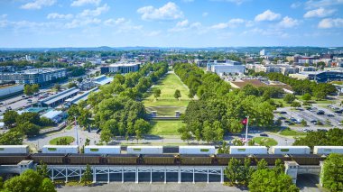 Aerial view of Nashville Bicentennial Capitol Mall State Park, lush greenery meets urban landscape. A train crosses the foreground under a bright sky, showcasing harmonious urban planning. clipart