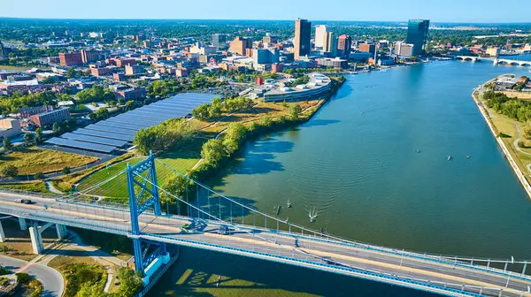 Toledo, Ohio 'nun canlı hava manzarası Maumee Nehri üzerindeki ikonik mavi Anthony Wayne Köprüsü' nü gösteriyor. Şehir merkezi ufuk çizgisi ve güneş panelleri kentsel büyüme ve sürdürülebilirliği yansıtıyor.