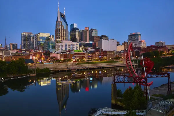 stock image Nashville vibrant skyline at dusk, with the iconic Batman Building, reflects in the Cumberland River. A red sculpture adds artistic flair, capturing the citys dynamic blend of architecture and