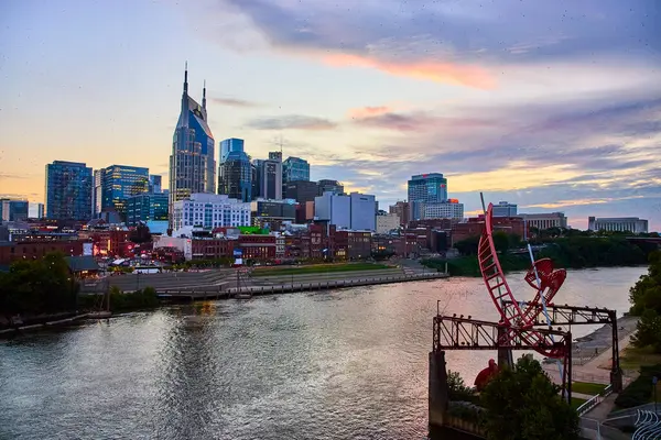 stock image Downtown Nashville skyline glows at sunset with the iconic Batman Building and Ghost Ballet sculpture by the Cumberland River. Experience the vibrant blend of urban art and cultural heritage.