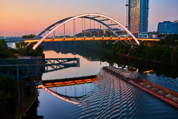 stock image Serene Nashville cityscape during blue hour showcasing the illuminated Korean Veterans Memorial Bridge reflected on the calm river as a barge passes by, highlighting urban charm and river commerce.