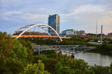 Serene Nashville akşamı: Kore Gaziler Anıtı köprüsü zemin zemininde pırıl pırıl parlayan bir gökdelenle bereketli nehir kıyıları üzerinde zarafetle kemer oluşturur. Şehir zarafeti doğal sükunetle buluşur..