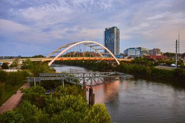 Nashville 'in bir bakışı: Koreli Gaziler Anıt Köprüsü, yemyeşil ve altın bir gökyüzü altında modern gökdelenlerin ufuk çizgisi tarafından çerçevelenmiş sakin bir nehrin üzerinde zarafetle kavis çiziyor..