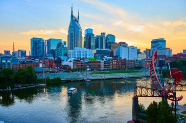 Altın Saatteki Nashville ufuk çizgisi: Batman Binası olarak bilinen ATT Binası, Cumberland Nehri üzerindeki kuleler, su üzerinde yansımalar ve ön planda canlı kırmızı bir heykel.