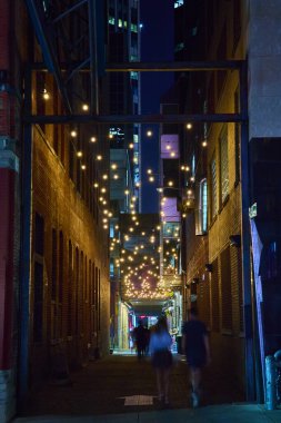 Sıcak sicim ışıkları Nashville Matbaa Sokağı 'nı gece aydınlatır. Uzun tuğlalı binalar ve bulanık figürler bu davetkar şehirde şehir cazibesini ve rahat gece hayatını çağrıştırıyor..