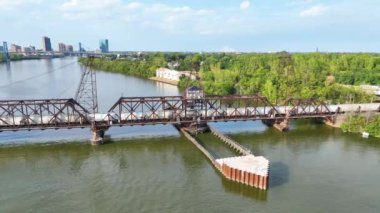 Aerial Arc Shot: Toledo endüstriyel cazibesini sakin bir nehrin üzerinde bir tren köprüsü ile keşfet. Kentsel ufuk çizgisi berrak gökyüzünün altında yemyeşil ile buluşuyor, mükemmel bir kentsel doğa karışımını somutlaştırıyor.