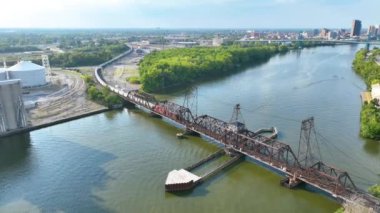 Toledo, Ohio 'nun Havadan Çekimi: Görkemli tren yolu, yemyeşil ve endüstriyel siloların ortasındaki sakin bir nehir boyunca uzanır ve sıcak öğleden sonra ışığı altında canlı şehir merkezinin siluetine karşı kurulur..