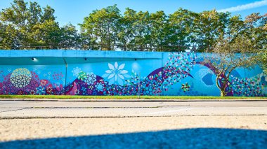 Fort Wayne Indiana 'daki canlı duvar resminde geometrik desenler ve kalın renklerde merkezi beyaz bir çiçek sergileniyor. Yeşilliğe karşı bir köprünün altında ele geçirilen bu sanat eseri kentsel yaratıcılığı kutluyor..