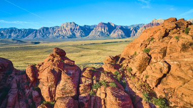Nevada 'daki Red Rock Kanyonu' nun altın saatindeki havadan görüntüsü. Canlı kırmızı kumtaşı oluşumları, geniş çöl havzasıyla ve parlak mavi gökyüzünün altındaki uzak dağlarla tezat oluşturuyor..