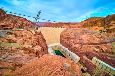 Aerial view of Hoover Dam, an engineering marvel nestled in the Black Canyon between Nevada and Arizona, showcasing its grandeur amid rugged terrain and a clear blue sky, with power lines spanning the clipart