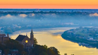 Cincinnati 'deki Ohio Nehri üzerinde tarihi bir kilise ve parlayan bir pencereyle sakin bir gündoğumunun havadan görüntüsü. Sis şehrin üzerinde süzülüyor altın saatinde dingin ve ruhani bir atmosfer yaratıyor..