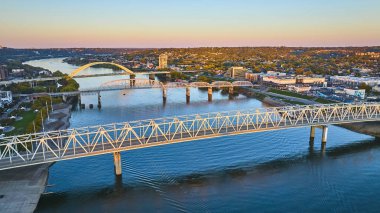 Aerial view of Cincinnati at golden hour, showcasing the iconic Daniel Carter Beard Bridge and Purple People Bridge over the Ohio River. The serene cityscape blends urban life with lush greenery. clipart