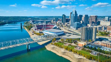 Cincinnati şehir merkezinin havadan görünüşü Great American Ball Park ve Paul Brown Stadyumu 'nu gösteriyor. Ohio Nehri ve ikonik Roebling Askılık Köprüsü, şehrin parlak siluetini ve spor haberlerini vurguluyor.